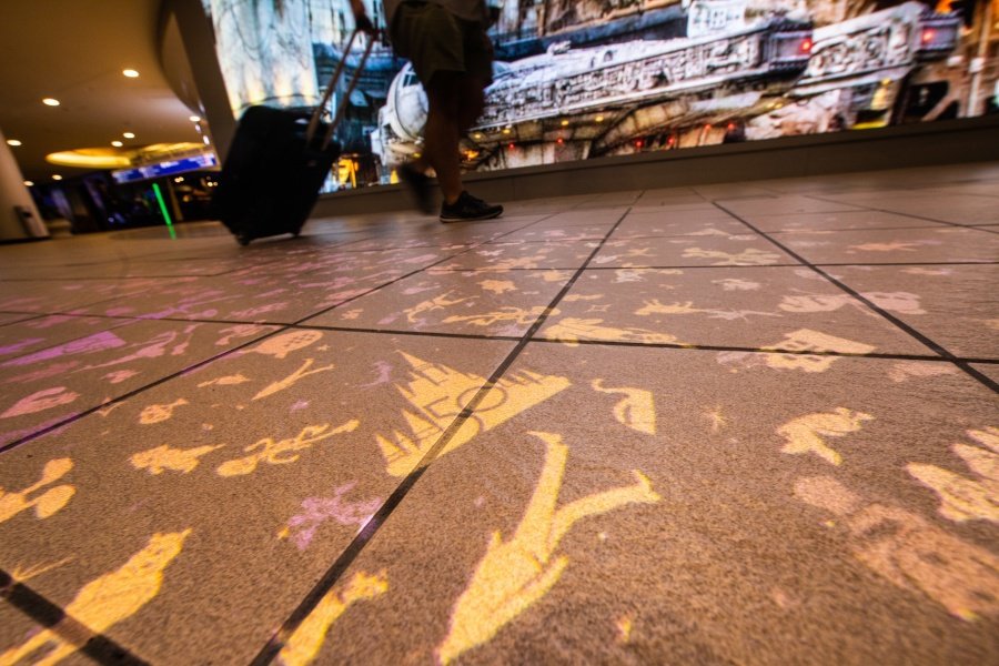 Silhouettes glide across the sunlit tile floor as families and fans drag their Disney-themed suitcases past a vibrant mural, capturing the rush of excitement that fills Orlando International Airport—the first step into the magic of Walt Disney World and nearby theme park adventures.