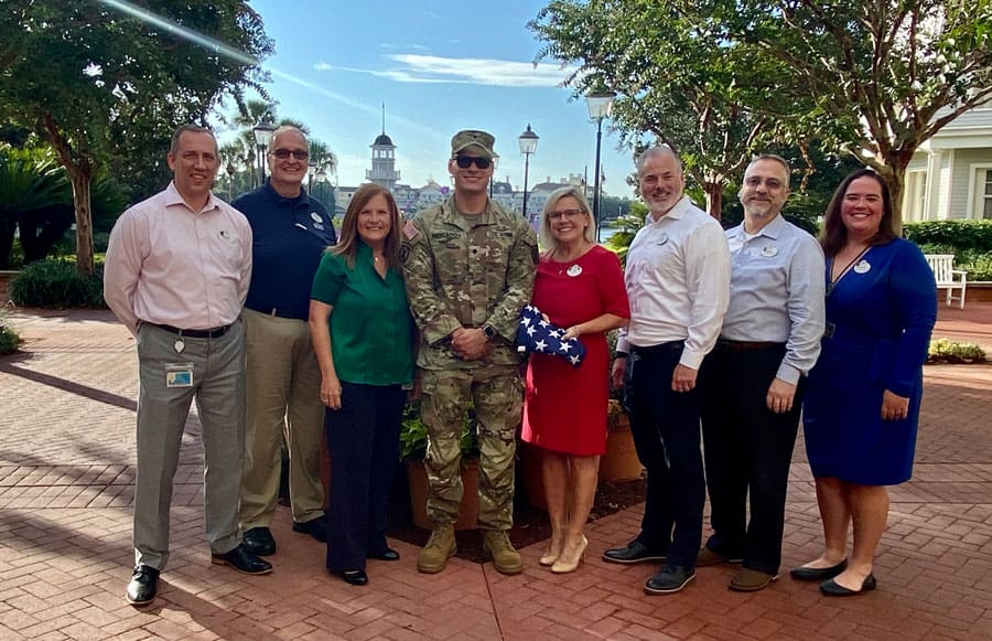 A group of eight guests, including a uniformed soldier, gather with joyful smiles beneath the bright sun at Disney World, celebrating a heartwarming hero’s return.