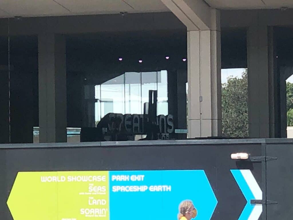 Construction walls with vibrant directional signs guide guests near EPCOT’s Creations Shop, while a fresh sign and shimmering building reflections in the background tease the exciting future updates coming to the park.