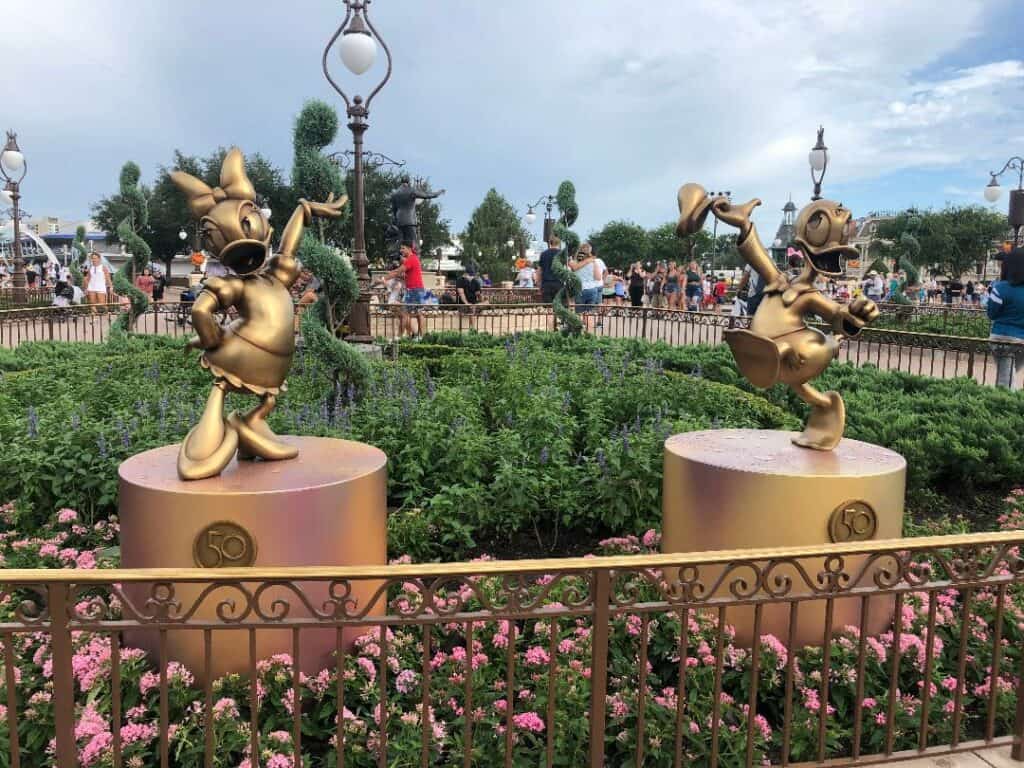 Daisy Duck and Donald Duck gleam in gold as Fab 50 Character Collection statues, showcased on pedestals amid blooming flowers at Magic Kingdom.