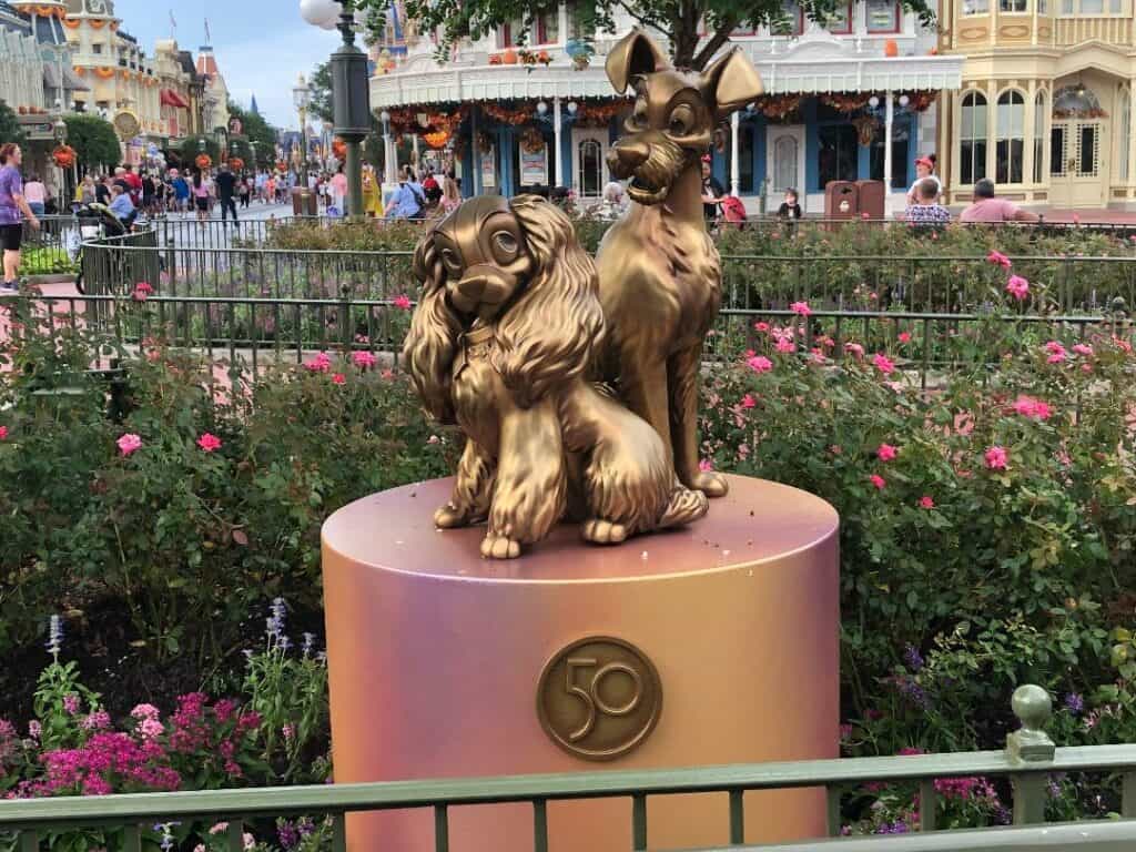 Lady and the Tramp bronze statue displayed on a pink pedestal featuring the 50th-anniversary emblem, surrounded by colorful flowers, as part of the Fab 50 Character Collection at Magic Kingdom.