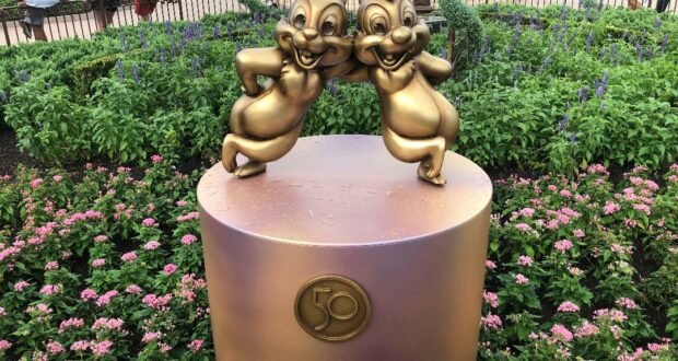 Chip and Dale’s gleaming gold statues from the Fab 50 Character Collection stand proudly on a round pedestal featuring a “50” emblem, surrounded by vibrant flowers in Magic Kingdom.
