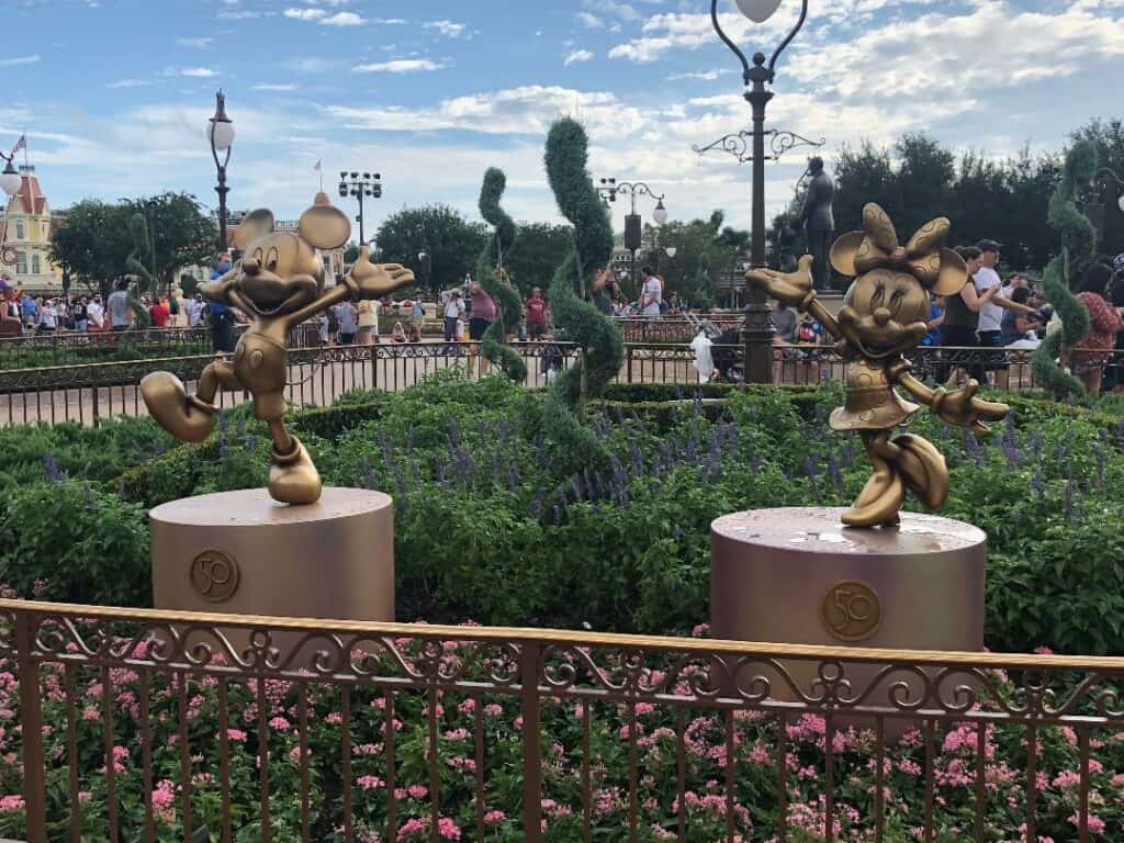 Golden Mickey and Minnie Mouse statues from the Fab 50 Character Collection shine brightly among vibrant flowers at Magic Kingdom, with guests enjoying the magical atmosphere in the background.