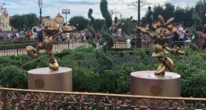 Golden Mickey and Minnie Mouse statues from the Fab 50 Character Collection shine brightly among vibrant flowers at Magic Kingdom, with guests enjoying the magical atmosphere in the background.