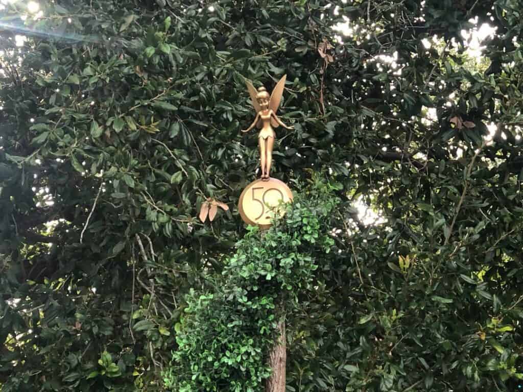 Tinker Bell from the Fab 50 Character Collection sparkles atop a golden "50" emblem, nestled among lush greenery, bringing extra enchantment to Magic Kingdom’s anniversary celebration.