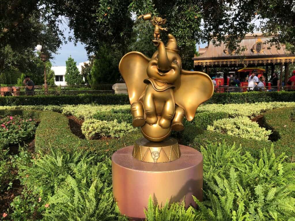 Dumbo from the Fab 50 Character Collection gleams in gold atop a pedestal, surrounded by lush garden greenery at Magic Kingdom.