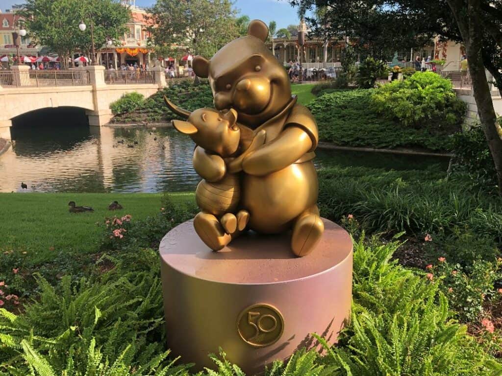 Winnie the Pooh and Piglet, featured in a shimmering golden Fab 50 Character Collection statue at Magic Kingdom, share a warm hug amid vibrant greenery and a picturesque pond backdrop.