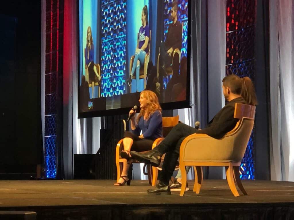 Panelists seated onstage at the Jodi Benson Panel at a fan convention, engaging in discussion as their conversation is broadcast above them on a giant screen for attendees.