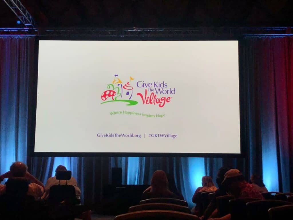 Theme park fans gather in an event space, watching a dynamic presentation featuring the Give Kids The World Village logo projected on a massive screen—celebrating the vital role this beloved charity plays within the theme park and entertainment community.