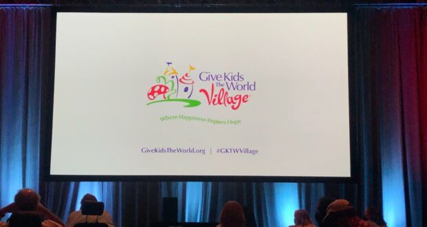 Theme park fans gather in an event space, watching a dynamic presentation featuring the Give Kids The World Village logo projected on a massive screen—celebrating the vital role this beloved charity plays within the theme park and entertainment community.
