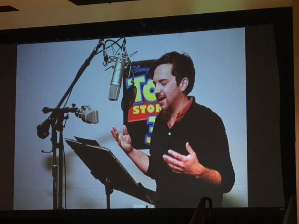 A voice actor performs at a microphone, with a Toy Story 3 poster on the wall behind him—just like John Morris's iconic vocal work celebrated at Disney and Pixar fan events.