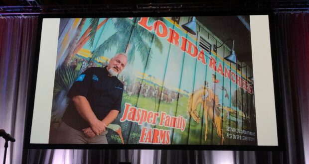 A guest gets ready to join a panel with Chef Art Smith onstage at an event, standing in front of a vibrant mural featuring the "Florida Ranches Jasper Family Farms" logo displayed across a large screen.