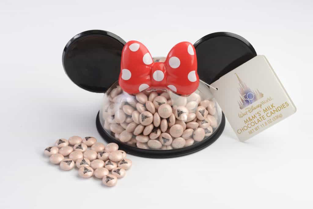 Minnie Mouse-themed candy container with her signature red bow, overflowing with pastel pink M&M’s and a whimsical tag attached. The sweet treats spill out just like the irresistible confections you’ll spot inside Main Street Confectionery at Magic Kingdom Park during its grand reopening.