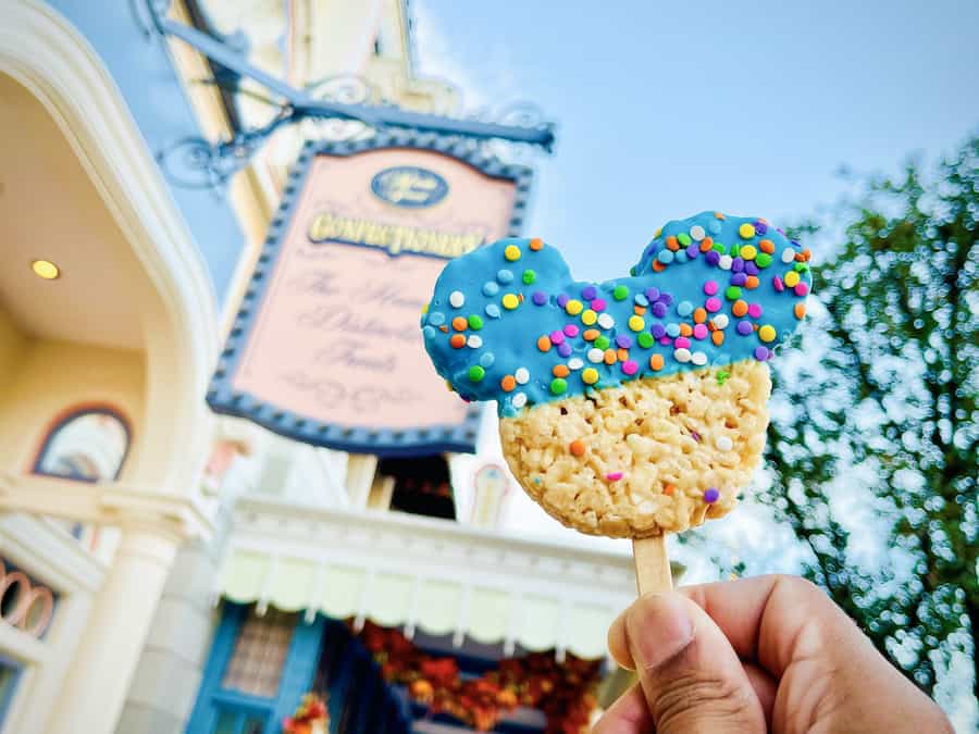 A Mickey-shaped Rice Krispies treat covered in blue icing and colorful sprinkles is held up in front of the freshly reopened Main Street Confectionery at Magic Kingdom, ready to delight Disney fans.