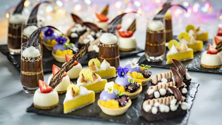 A tempting display of Disney desserts—chocolate treats, fruity sweets, and edible flowers—beautifully presented on sleek black trays, ready for Dessert Parties. Vibrant, blurred lights in the background evoke Disney Enchantment magic, making this spread perfect for guests celebrating the fireworks spectacular on October 3rd.