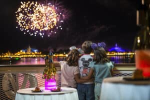 Three kids stand side by side at the edge of a lake in Magic Kingdom, gazing up at breathtaking Disney Enchantment fireworks illuminating the night sky, fully immersed in the wonder and excitement of the show.