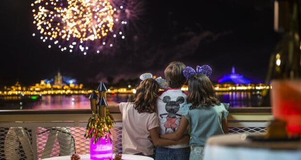 Three kids stand side by side at the edge of a lake in Magic Kingdom, gazing up at breathtaking Disney Enchantment fireworks illuminating the night sky, fully immersed in the wonder and excitement of the show.