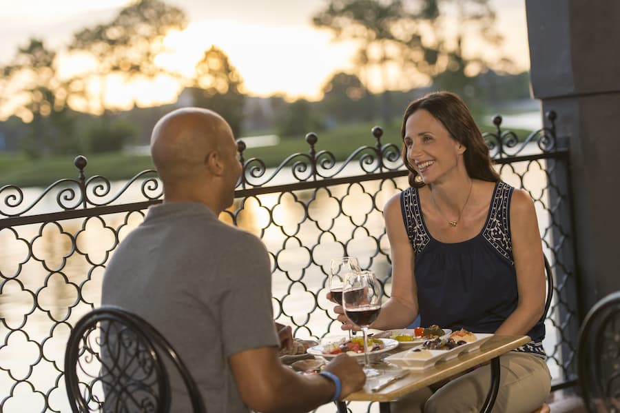 A couple relaxes at an outdoor dining area in a theme park at sunset, laughing and chatting over plates of food and glasses of red wine, soaking up the festive atmosphere and discovering fresh ways to connect amid lively attractions.