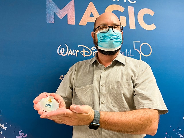 Jose, wearing a mask, proudly holds his Cast Member badge in front of the Walt Disney World 50th anniversary backdrop, celebrating the incredible team members who bring the Disney magic to life every day.