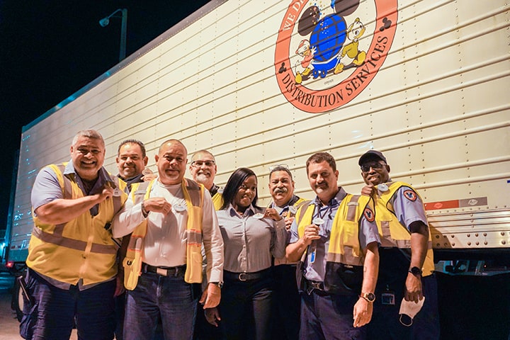 Nine Disney Cast Members in their signature work uniforms smile and pose together in front of a large Disney distribution truck, proudly celebrating the spirit of teamwork that keeps the magic alive behind the scenes.