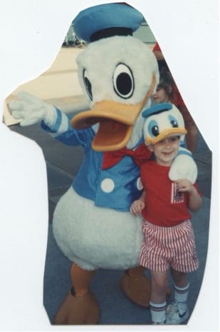 A young guest in a vibrant red outfit beams alongside Donald Duck, both wearing festive hats and sharing a joyful moment at Walt Disney World’s 50th Anniversary celebration.