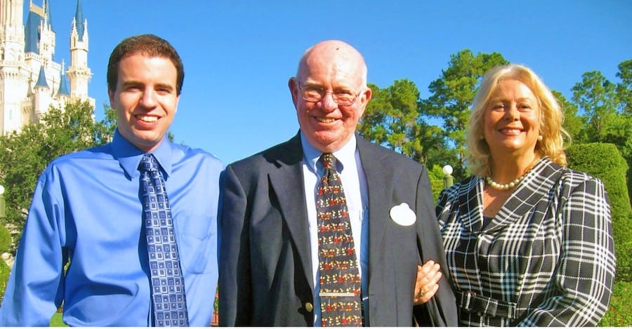 Three guests dressed in formalwear beam with excitement outside Cinderella Castle at Walt Disney World, surrounded by lush trees and sunshine, commemorating the resort’s 50th Anniversary celebration.