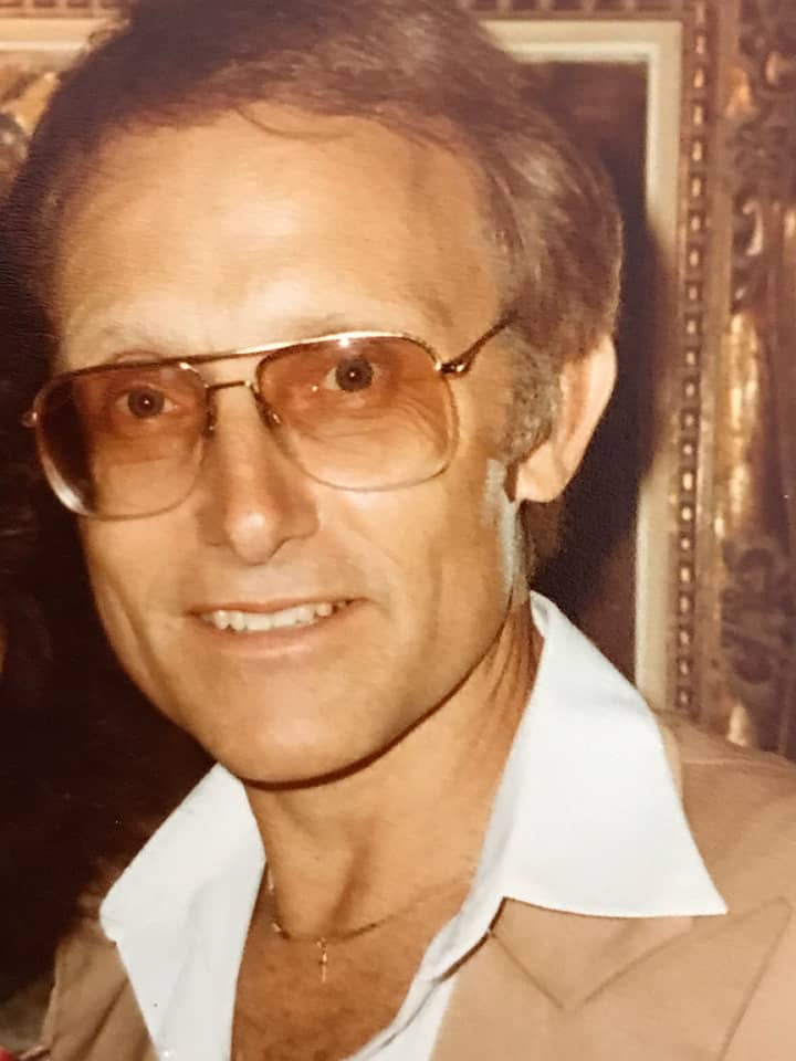 A proud Walt Disney World Cast Member with glasses, light brown hair, and a tan jacket beams indoors, framed by celebratory artwork as he marks 50 years of magical memories at the resort.
