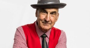 Yehaa Bob, wearing his signature red vest, patterned shirt, and gray hat, flashes a big smile in front of a plain white backdrop—showcasing the high-energy fun of his Modified Show beloved by theme park fans.