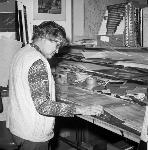 Inspiring nod to Disney legend Ruthie Tompson: an older woman in a patterned sweater, immersed in her work at a desk overflowing with animation artwork and storyboards, honoring the legacy of the iconic Disney animator who shaped so many classic films and whose remarkable life spanned 111 years.