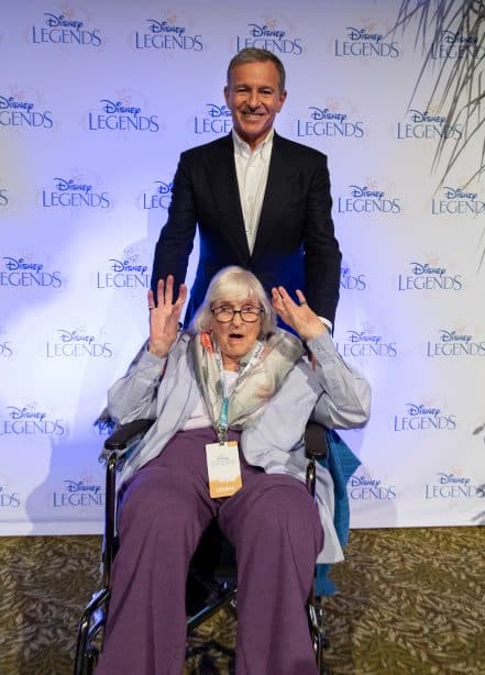 Ruthie Tompson beams and waves from her wheelchair as a man in a suit stands proudly behind her during the Disney Legends ceremony, where fans and Disney cast members honor her extraordinary contributions to animation before she passes away at 111.