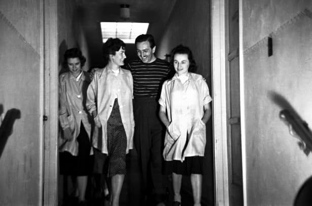 Three women and one man stroll down a Disney studio hallway, their faces lit with smiles as they chat animatedly—likely sharing fond memories of legendary animator Ruthie Tompson, whose creative legacy at Disney continues to inspire fans and cast members even after her passing at 111.