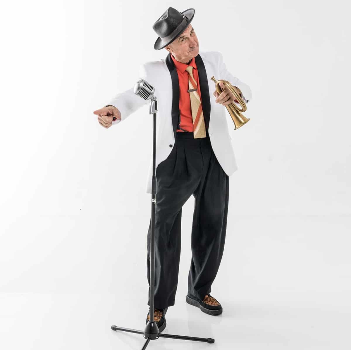 A charismatic performer in a crisp white jacket and hat stands beside a microphone, trumpet in hand, pointing enthusiastically to the crowd against a bright backdrop—channeling Yehaa Bob’s dynamic energy and inviting guests to experience the modified show’s lively entertainment.