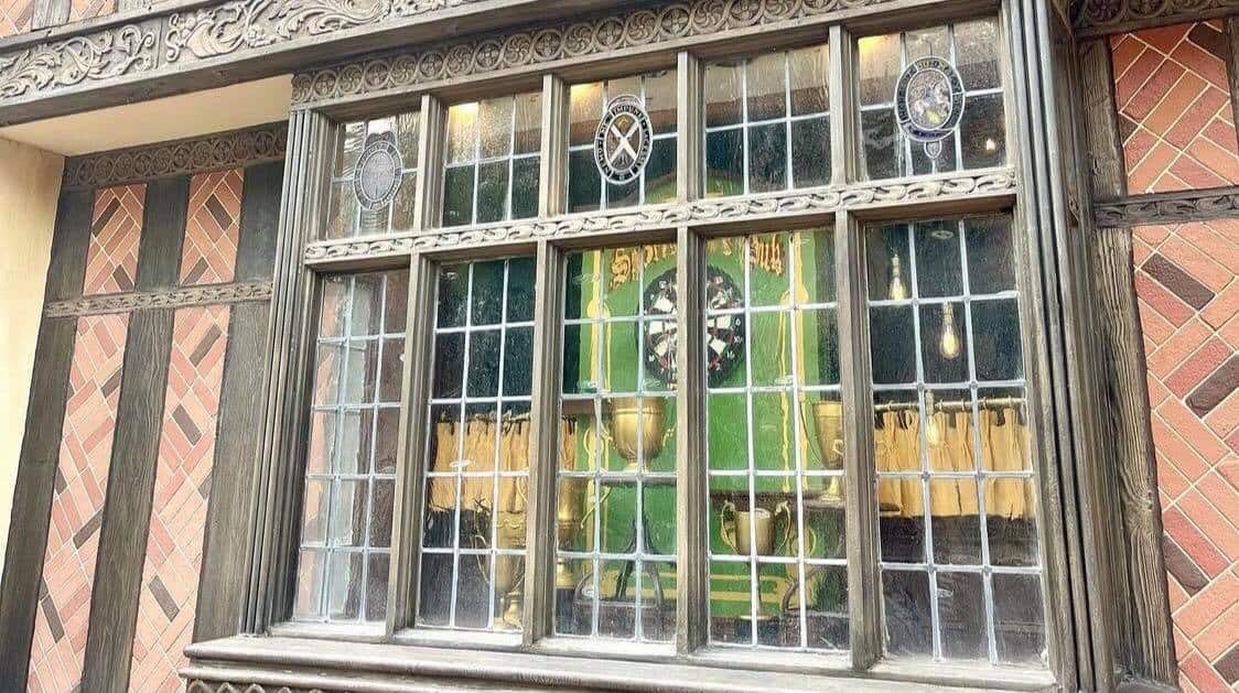A beautifully crafted vintage window with intricate glasswork showcases a vibrant green display and gleaming gold accents inside—an enchanting new feature that captures the classic charm of EPCOT’s United Kingdom Pavilion.