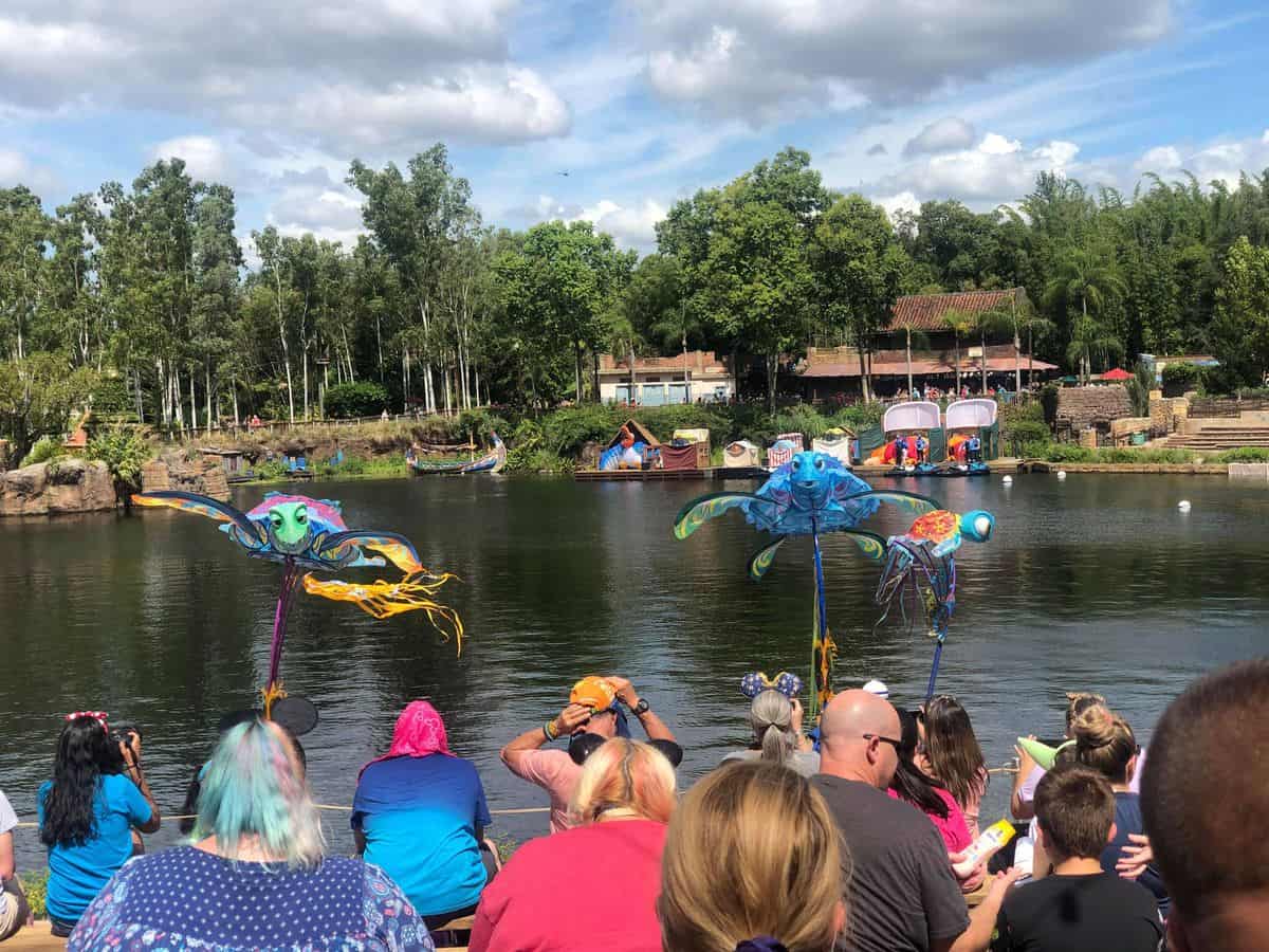 Guests at Disney’s Animal Kingdom enjoy the enchanting KiteTales show by Discovery River, watching vibrant fish puppets glide near the water as boats cruise past and lush trees frame the background beneath a partly cloudy sky, while colorful kites dance overhead in a celebration of Disney magic.