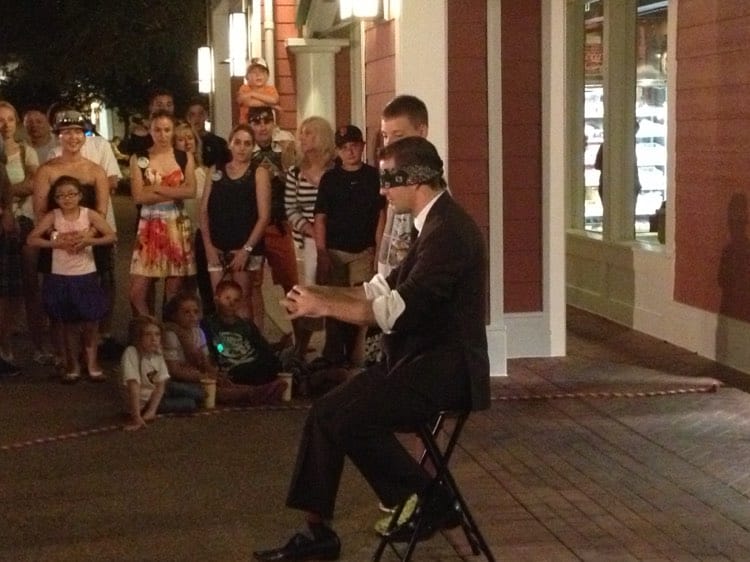 A magician, elegantly dressed and blindfolded, dazzles excited guests with a captivating street performance outside Jellyrolls on the BoardWalk at night, creating the same electric anticipation as waiting for a one-of-a-kind nighttime show at Walt Disney World.