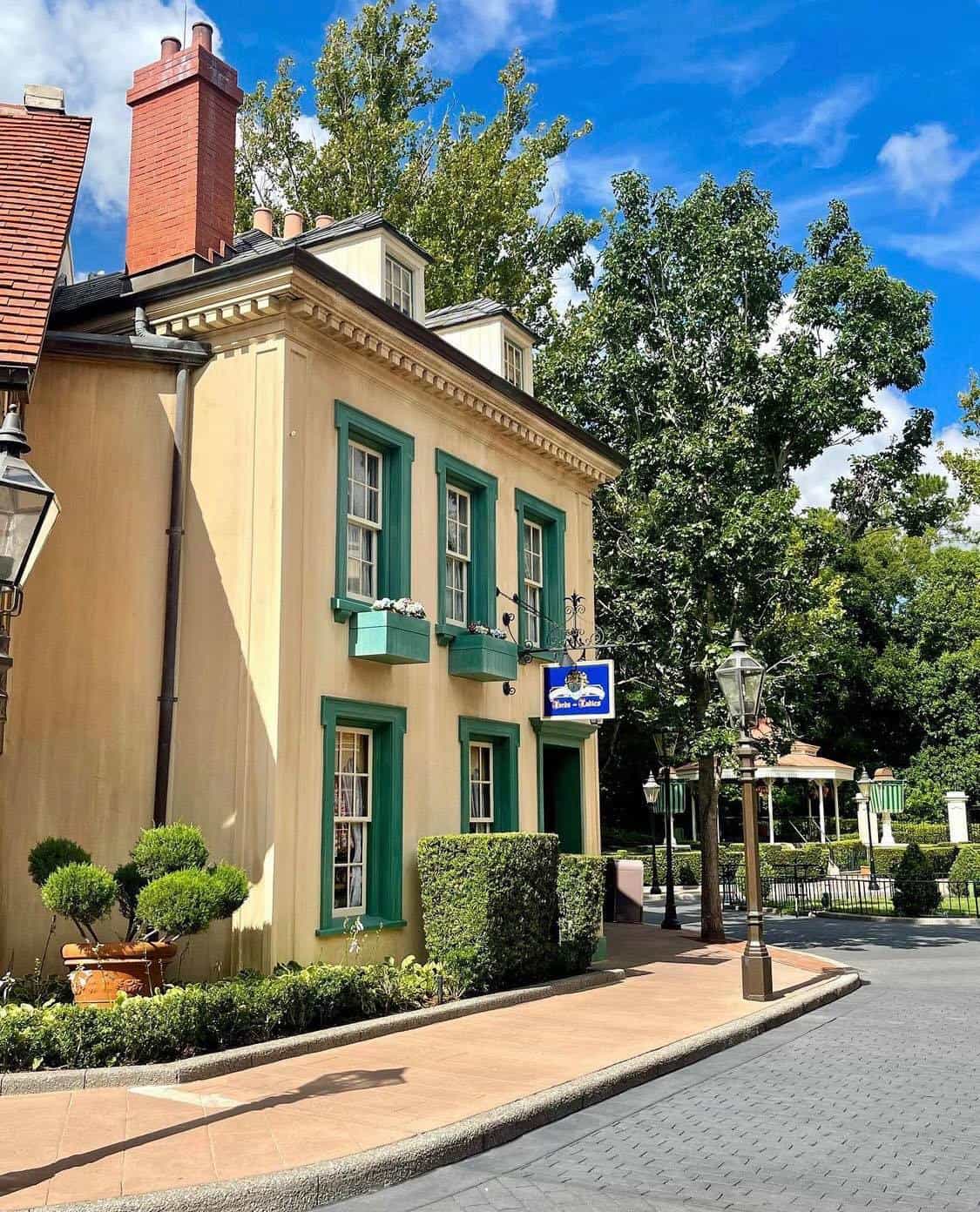 Charming two-story yellow building with classic green trim and lush landscaping, nestled among trees beneath a bright blue sky—authentically styled to capture the spirit of EPCOT’s United Kingdom Pavilion.