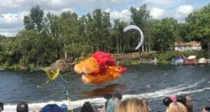 Simba-soaring kite takes flight over the Discovery River at Disney’s Animal Kingdom, capturing the awe of a crowd during KiteTails, with lush trees and vibrant blue sky setting the stage for this unforgettable Disney entertainment moment.