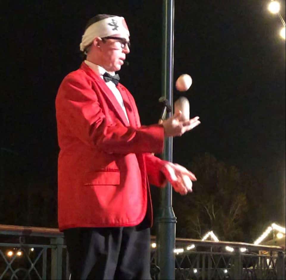 A flamboyantly dressed performer in a red jacket and bow tie dazzles guests with an energetic juggling act beneath twinkling string lights, kicking off the Opening Date celebration of Jellyrolls with lively entertainment.
