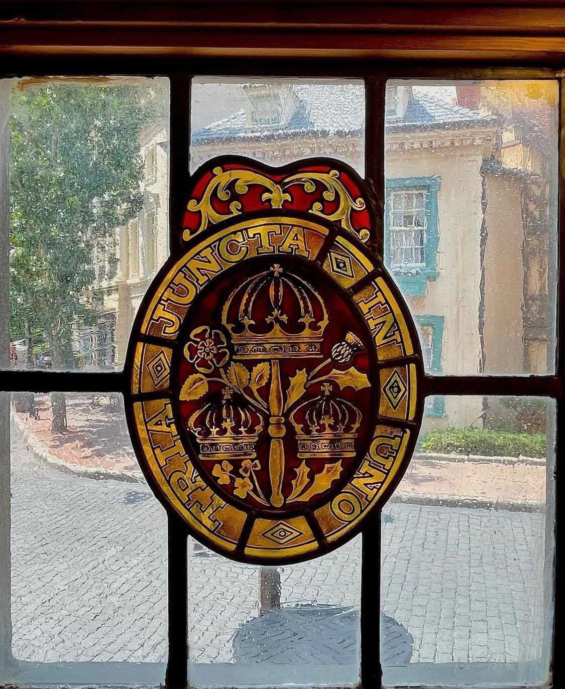 Elegant stained glass featuring crowns and the phrase "TRIA JUNCTA IN UNO" in gold shines above a scenic cobblestone street, adding royal flair to the United Kingdom Pavilion at EPCOT.