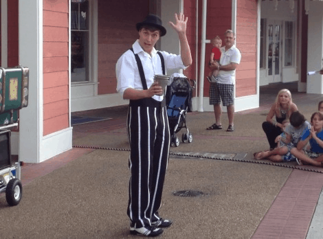 Guests seated outside the vibrant red Jellyrolls buildings watch as a lively street performer in striped pants waves and entertains, adding excitement to the crowd eagerly awaiting the Opening Date festivities.