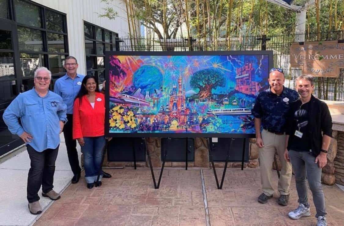 Five Disney fans gather around a vibrant painting celebrating Walt Disney World's 50th Anniversary, showcased on an easel outdoors in the park.