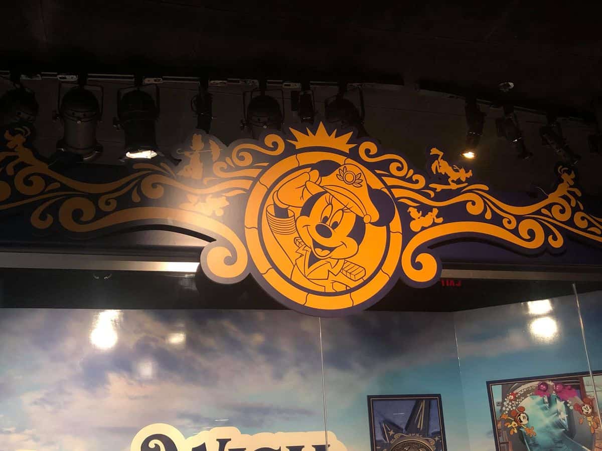 A golden Minnie Mouse captain's emblem shines with intricate detailing on a lavishly decorated wall, illuminated by theatrical lighting at Disney's Hollywood Studios.