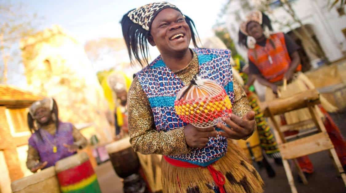 A Tam Tam Drummers performer in vibrant attire smiles while playing a shekere, with fellow drummers energizing the crowd at Harambe in Disney’s Animal Kingdom on November 7th.