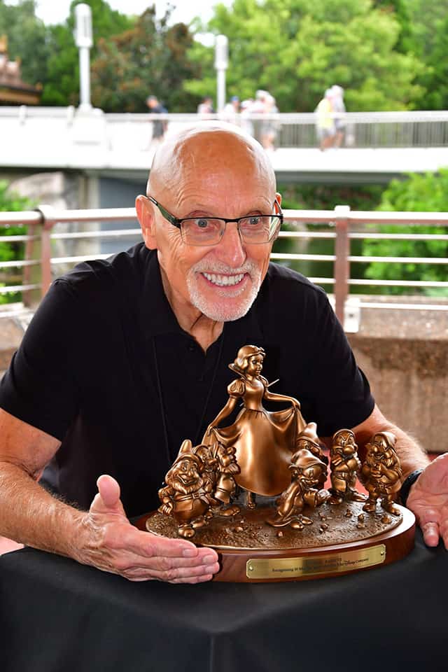 A Cast Member beams with pride, showing off a bronze Snow White and Seven Dwarfs sculpture on a black tablecloth as part of the 50 Years of Walt Disney World celebration.