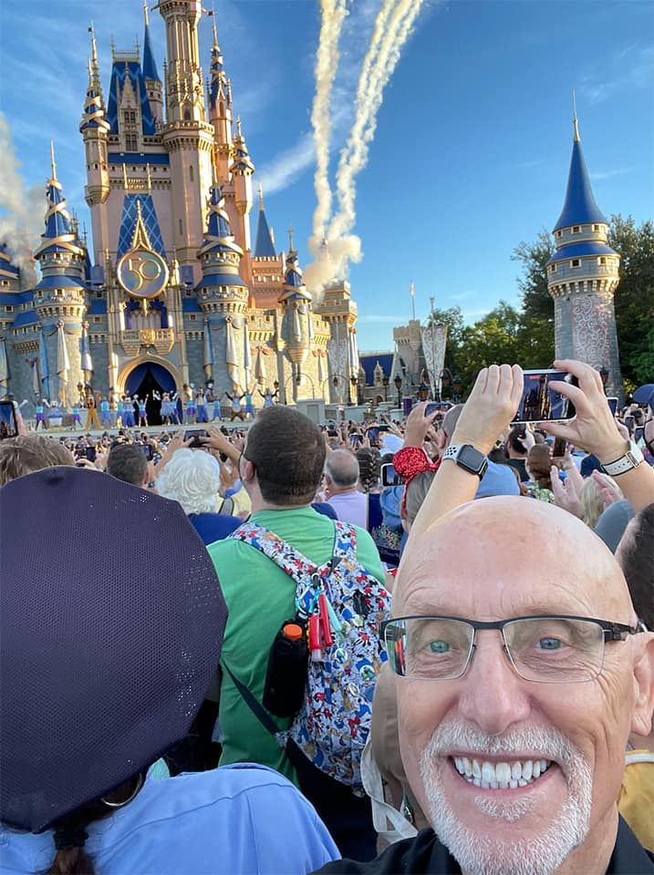 Guest shares a joyful selfie with a Walt Disney World Cast Member in front of Cinderella Castle, surrounded by excited crowds during the 50th Anniversary celebration, as vibrant fireworks light up the blue sky.