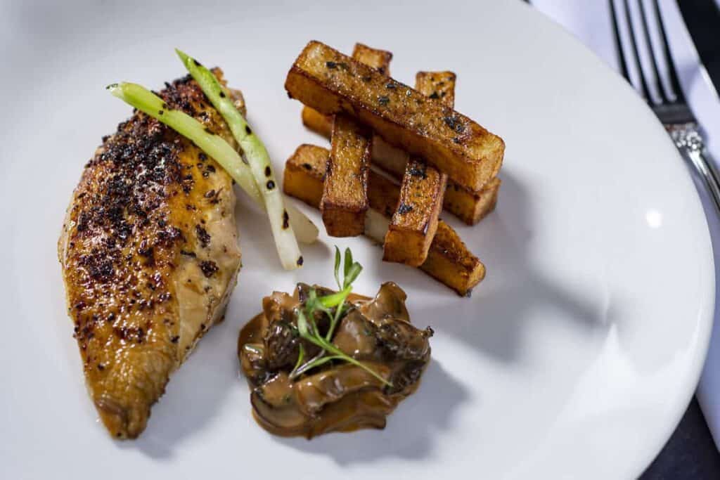 A plated entrée from the Frozen Dining Adventure aboard Disney Wish featuring grilled chicken breast, stacked fries, sautéed mushrooms, and green onions artfully arranged on a white plate—capturing the magic and culinary flair of Disney Cruise Line dining.
