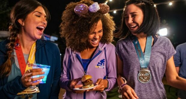 Three friends sporting runDisney medals share laughs late at night during the Disney Wine & Dine Post Race Party at EPCOT; one savors a glowing specialty drink while another enjoys a themed dessert plate, surrounded by the magical nighttime atmosphere.