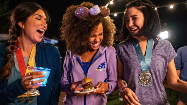 Three friends sporting runDisney medals share laughs late at night during the Disney Wine & Dine Post Race Party at EPCOT; one savors a glowing specialty drink while another enjoys a themed dessert plate, surrounded by the magical nighttime atmosphere.