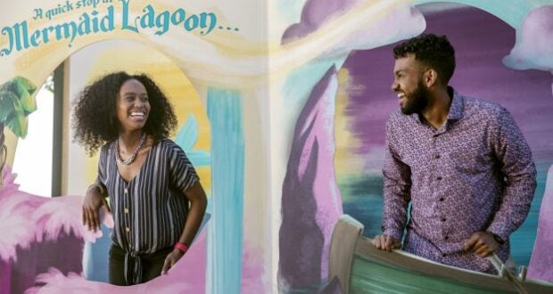 Guests share a joyful moment in front of the vibrant Mermaid Lagoon backdrop, featuring playful waves and whimsical clouds at EPCOT International Festival of the Arts.