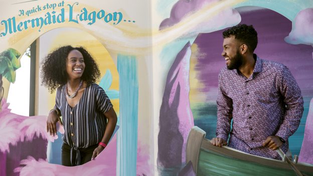 Guests share a joyful moment in front of the vibrant Mermaid Lagoon backdrop, featuring playful waves and whimsical clouds at EPCOT International Festival of the Arts.